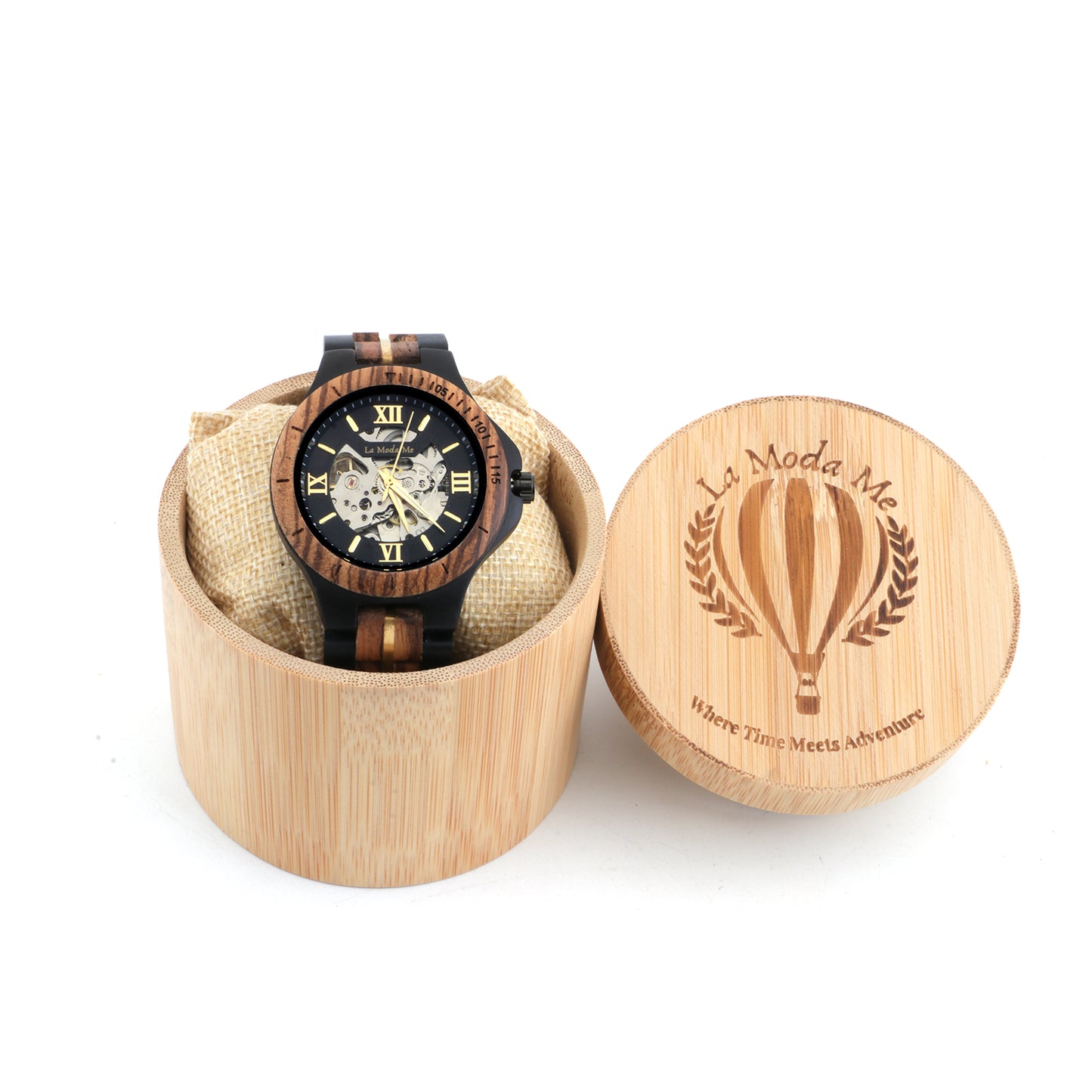 Chronograph wristwatch with a black and gold skeleton dial, displayed on a cushion inside a round bamboo gift box. The watch features a dark wood and metal link strap, and the box lid is engraved with the “La Moda Me” logo and the phrase “Where Time Meets Adventure”.
